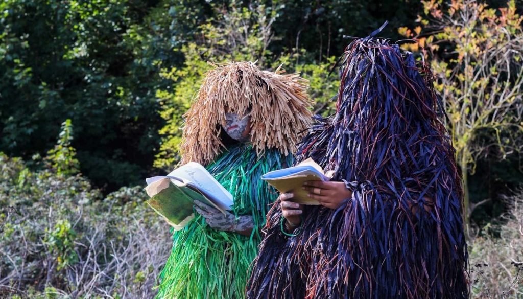 Two people wearing outfits made of recycled materials and mixed media reading books in the countryside