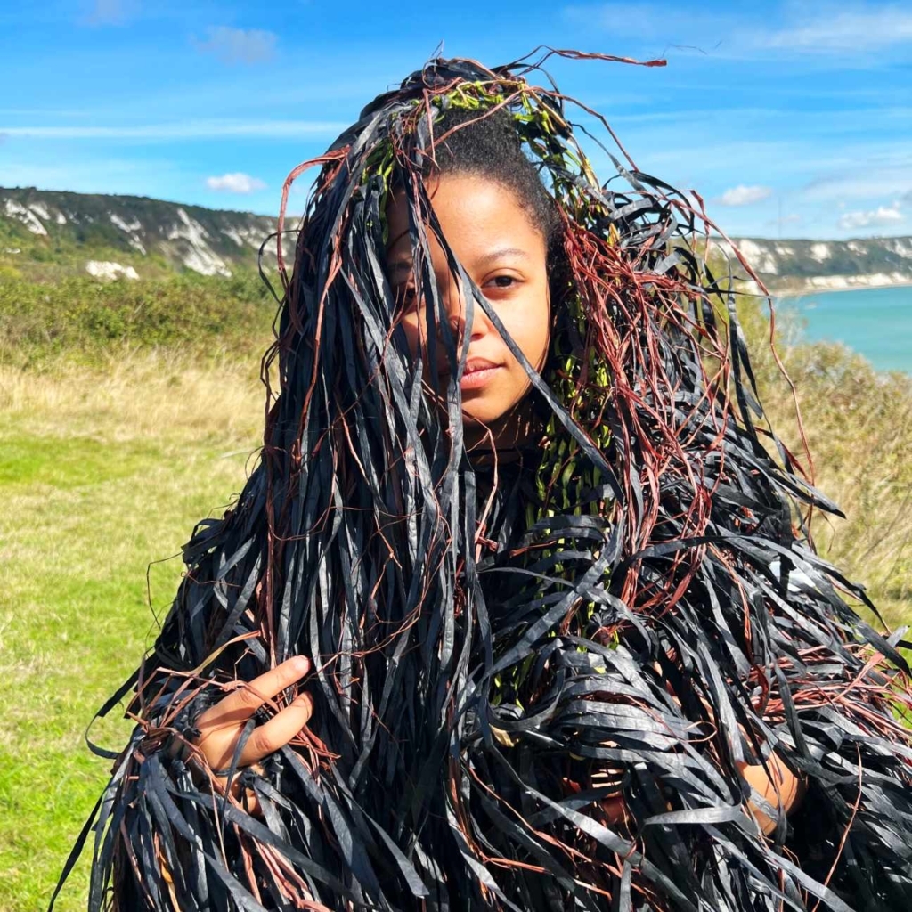Woman of colour wearing outfit made of recycled materials and mixed media against the backdrop of The Warren, Folkestone
